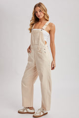 woman wearing beige overalls with adjustable straps and buttons, styled with a white tank top and sneakers