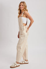 woman wearing a beige denim overall with a white tank top underneath and casual sneakers