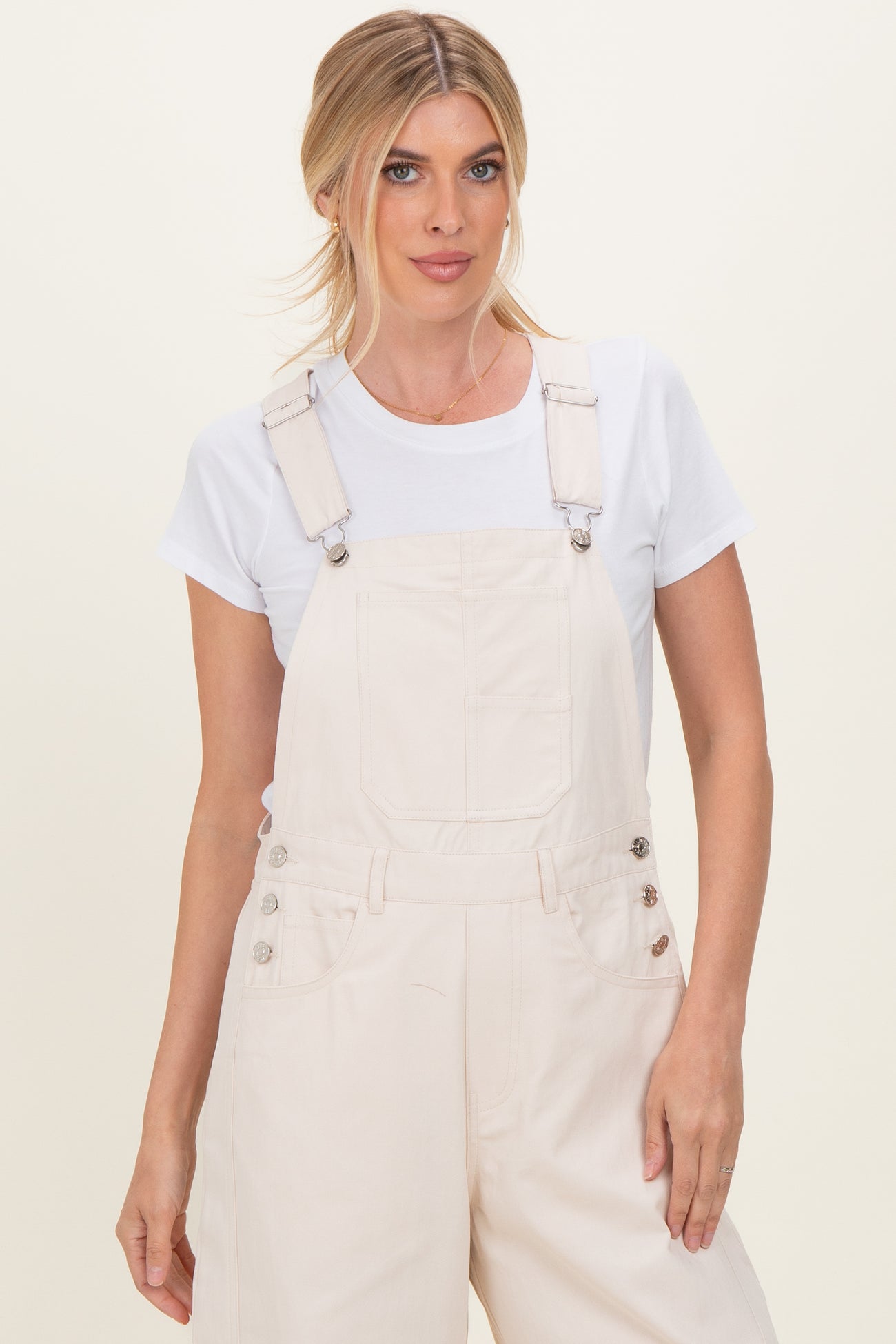woman wearing a white t-shirt underneath cream-colored denim overalls with adjustable straps and button details