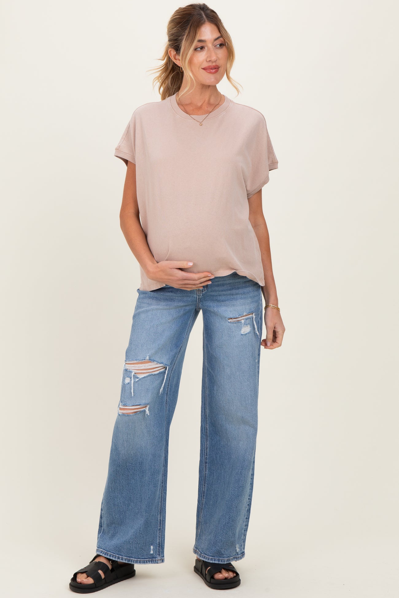 woman wearing a light pink maternity top and distressed wide-leg jeans, holding her baby bump