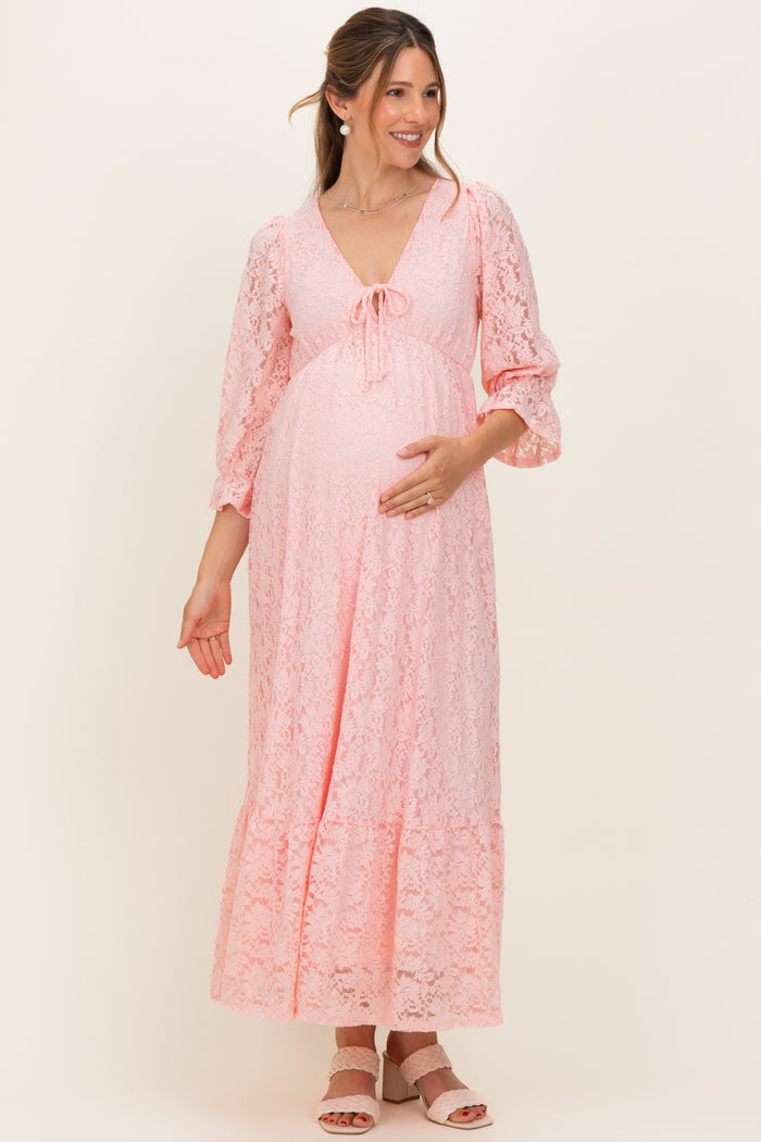 woman wearing a pink lace maternity maxi dress with three-quarter sleeves and a tie detail, holding her baby bump
