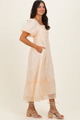 woman wearing a light orange gingham midi dress with puff sleeves and pockets, paired with strappy block heel sandals