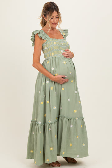 woman wearing a light green maternity maxi dress with floral embroidery and ruffled straps, cradling her baby bump