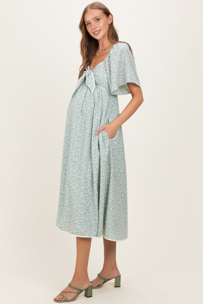 woman wearing a green floral print maternity dress with a bow detail and pockets, showing her baby bump