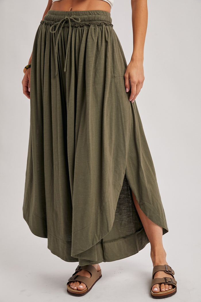woman wearing an olive green maxi skirt with a high slit and elastic waist, standing with one hand on her hip