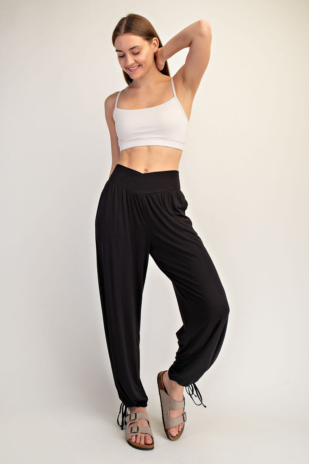 woman wearing a white crop top and black drawstring jogger pants, smiling and posing with one hand on her hip