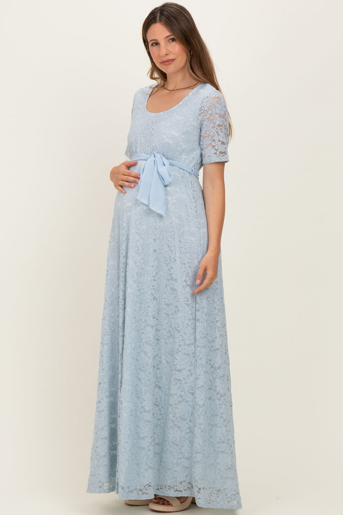 woman wearing a light blue lace maternity gown with a bow at the waist and holding her baby bump