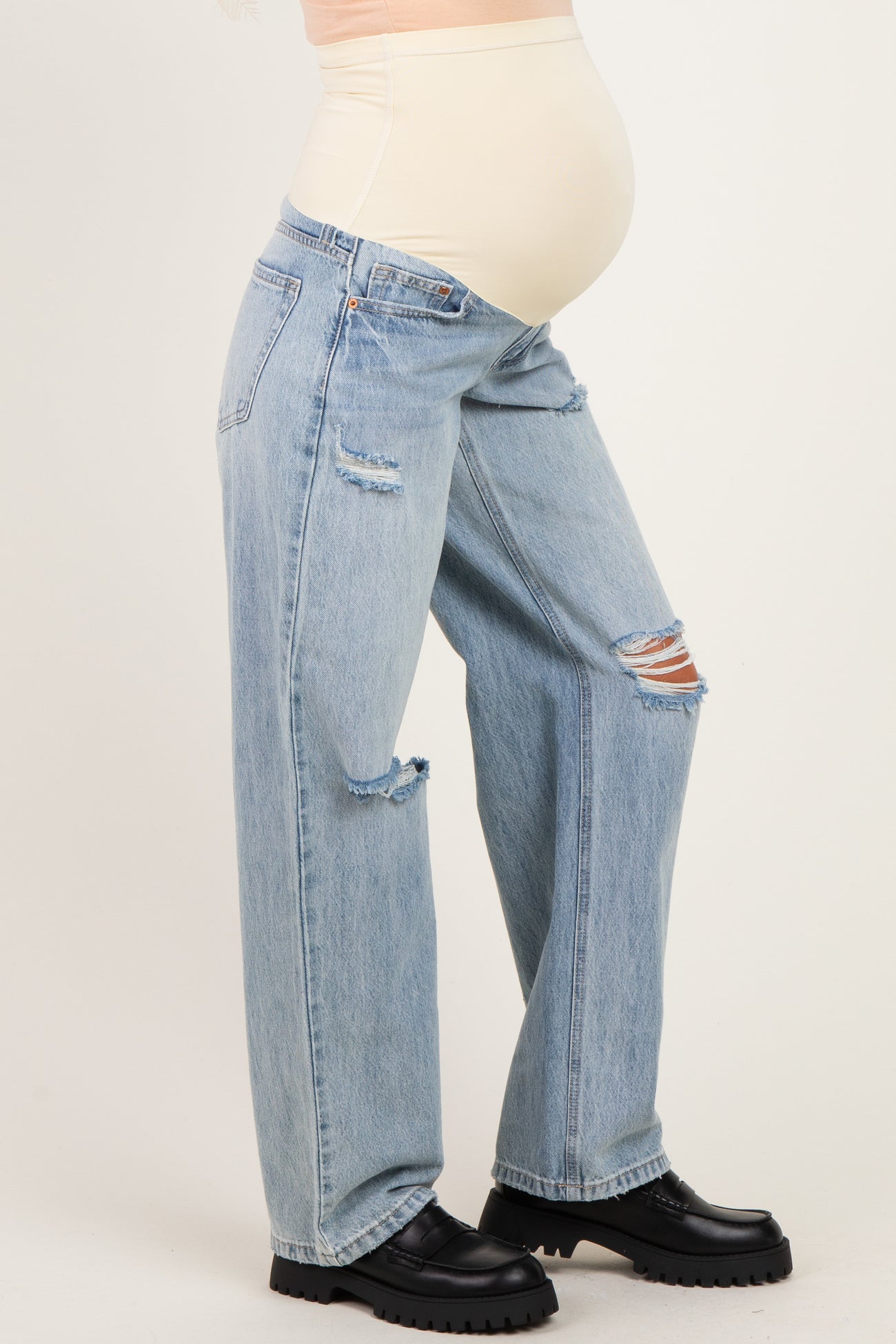 woman wearing light blue distressed maternity jeans and a beige belly support top, showcasing her baby bump