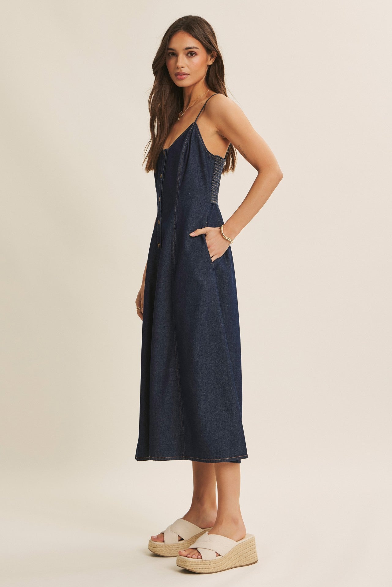 woman wearing a dark denim midi dress with adjustable straps and side pockets, paired with beige wedge sandals