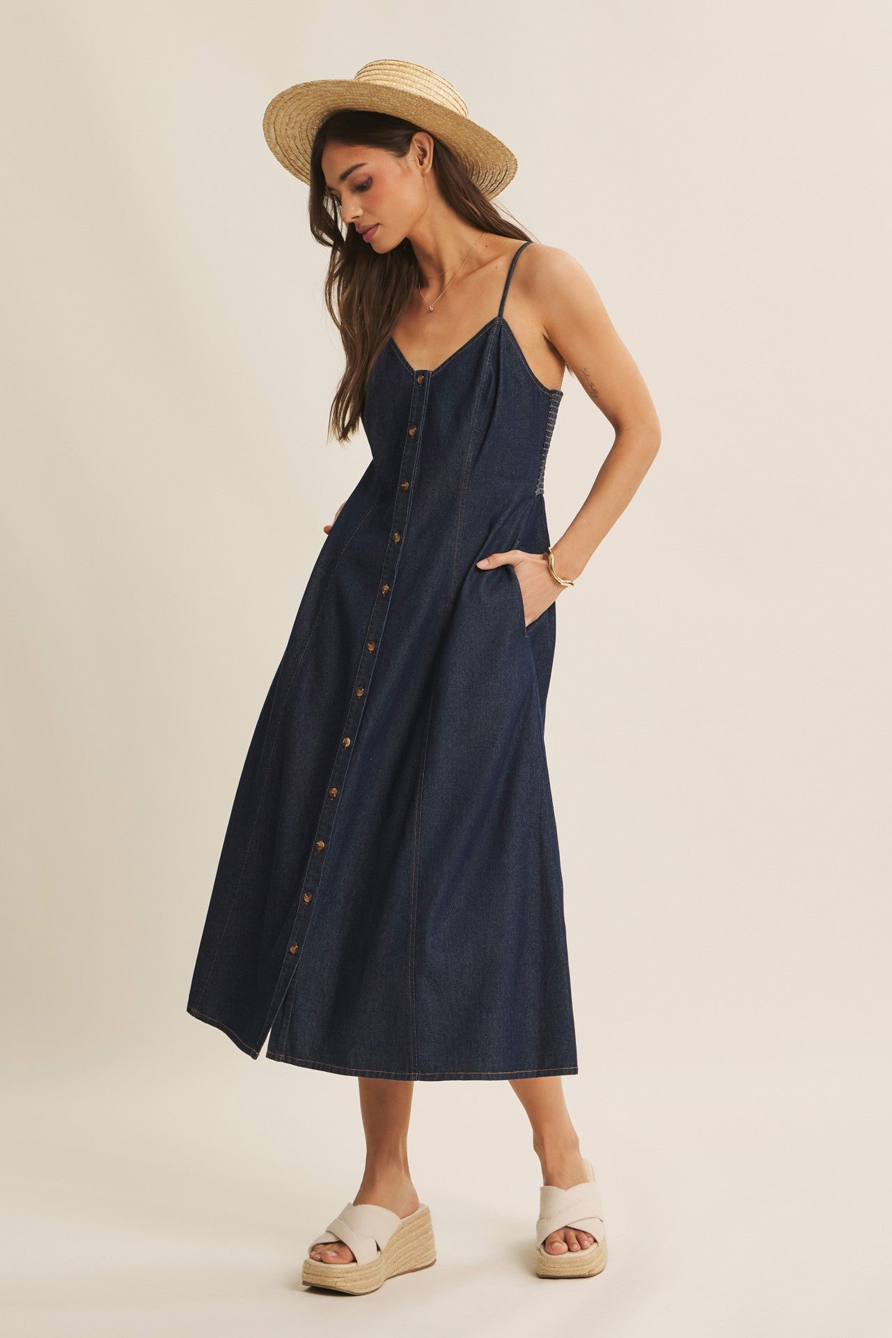 woman wearing a dark denim button-up maxi dress with thin straps and pockets, paired with a straw hat and wedge sandals