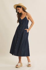 woman wearing a dark denim button-up maxi dress with thin straps and pockets, paired with a straw hat and wedge sandals