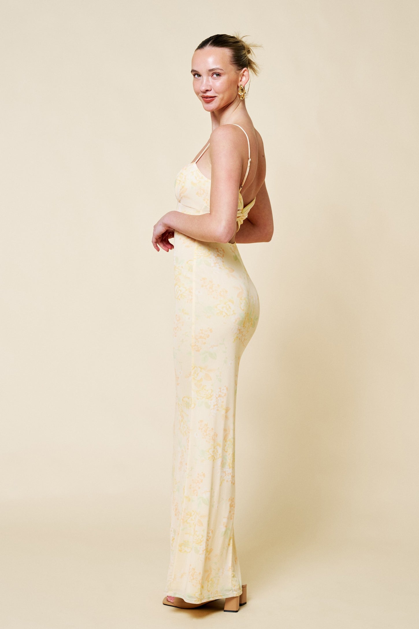 woman wearing a yellow floral spaghetti strap maxi dress, standing sideways with a soft smile and hair styled up