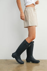woman wearing a white t-shirt, striped shorts, and black rain boots standing against a wall