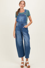 woman wearing a blue denim maternity overall with a green short sleeve top and standing with one hand in the pocket