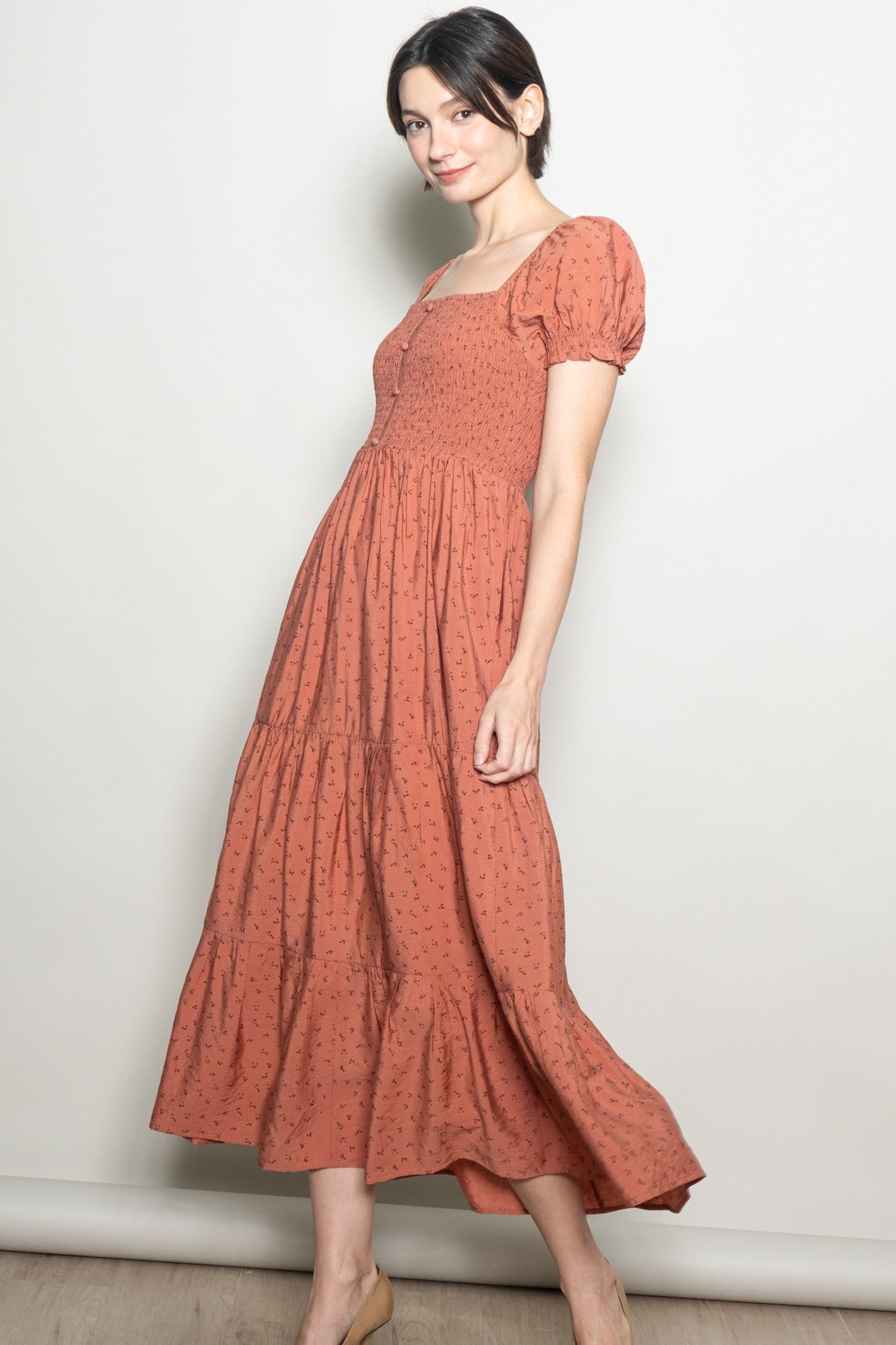 woman wearing a rust-colored midi dress with puff sleeves and a smocked bodice standing sideways against a neutral background