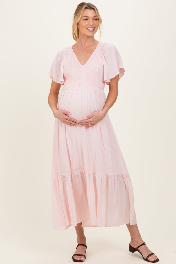 woman wearing a light pink maternity dress with flutter sleeves and a baby bump, standing and holding her belly