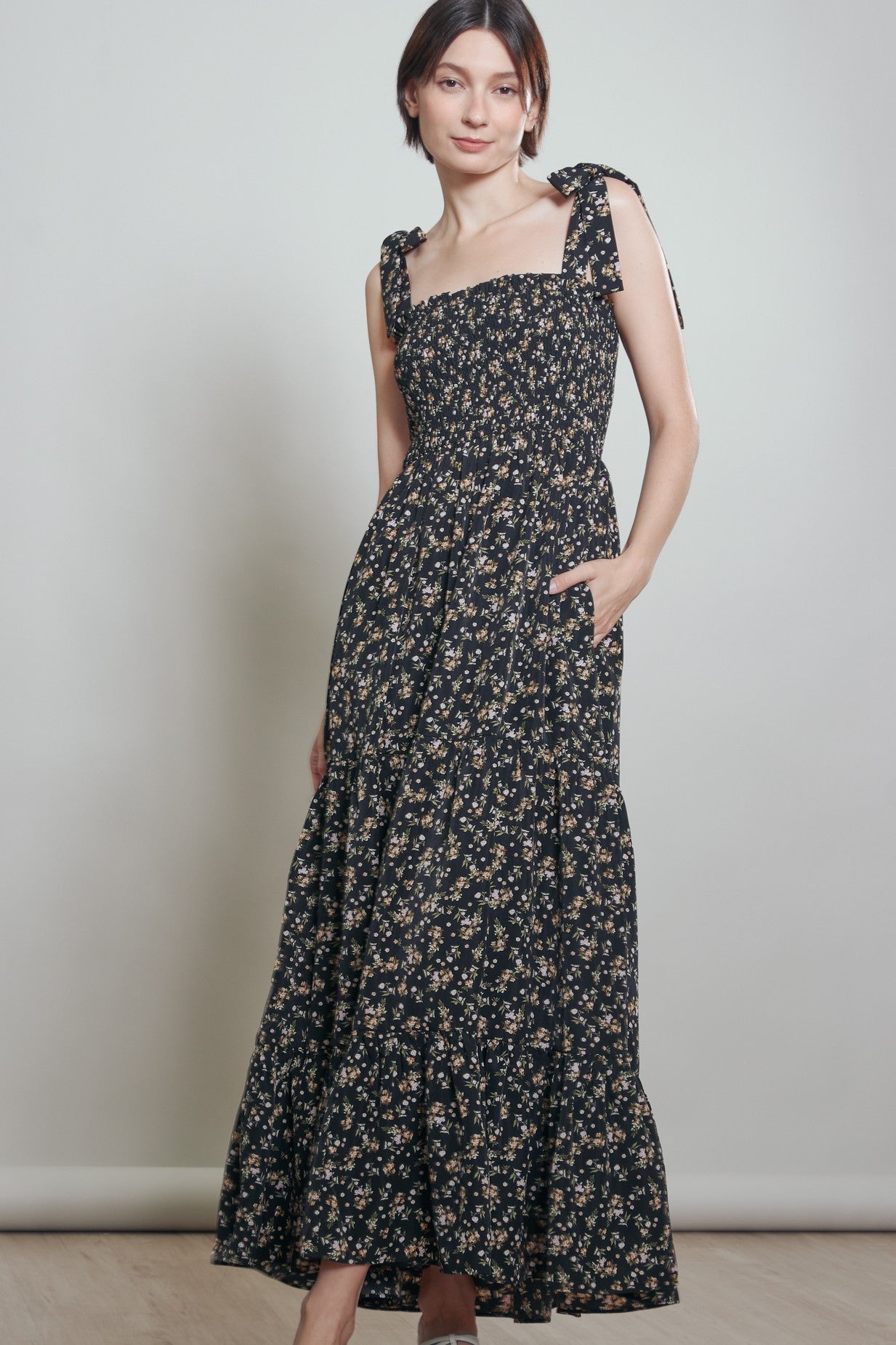woman wearing a black floral maxi dress with tie straps and pockets, standing against a neutral background