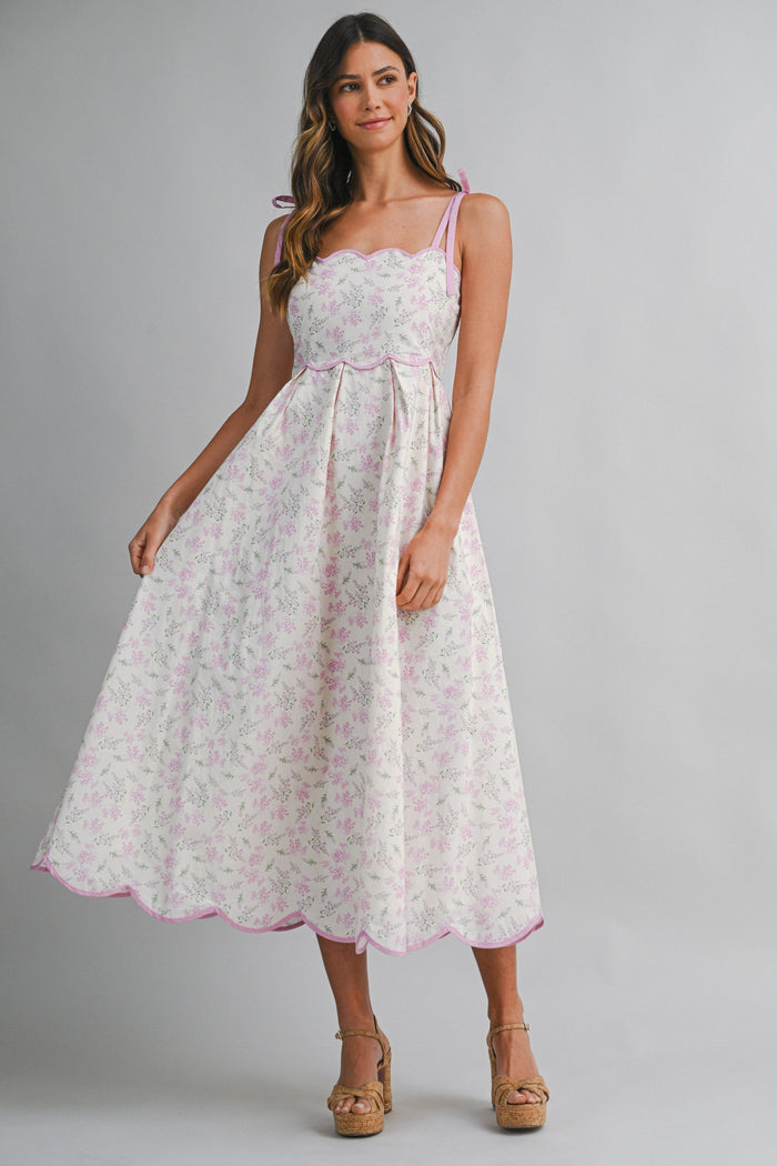 woman wearing a white floral midi dress with pink accents and tie straps, smiling and standing with one hand on her hip