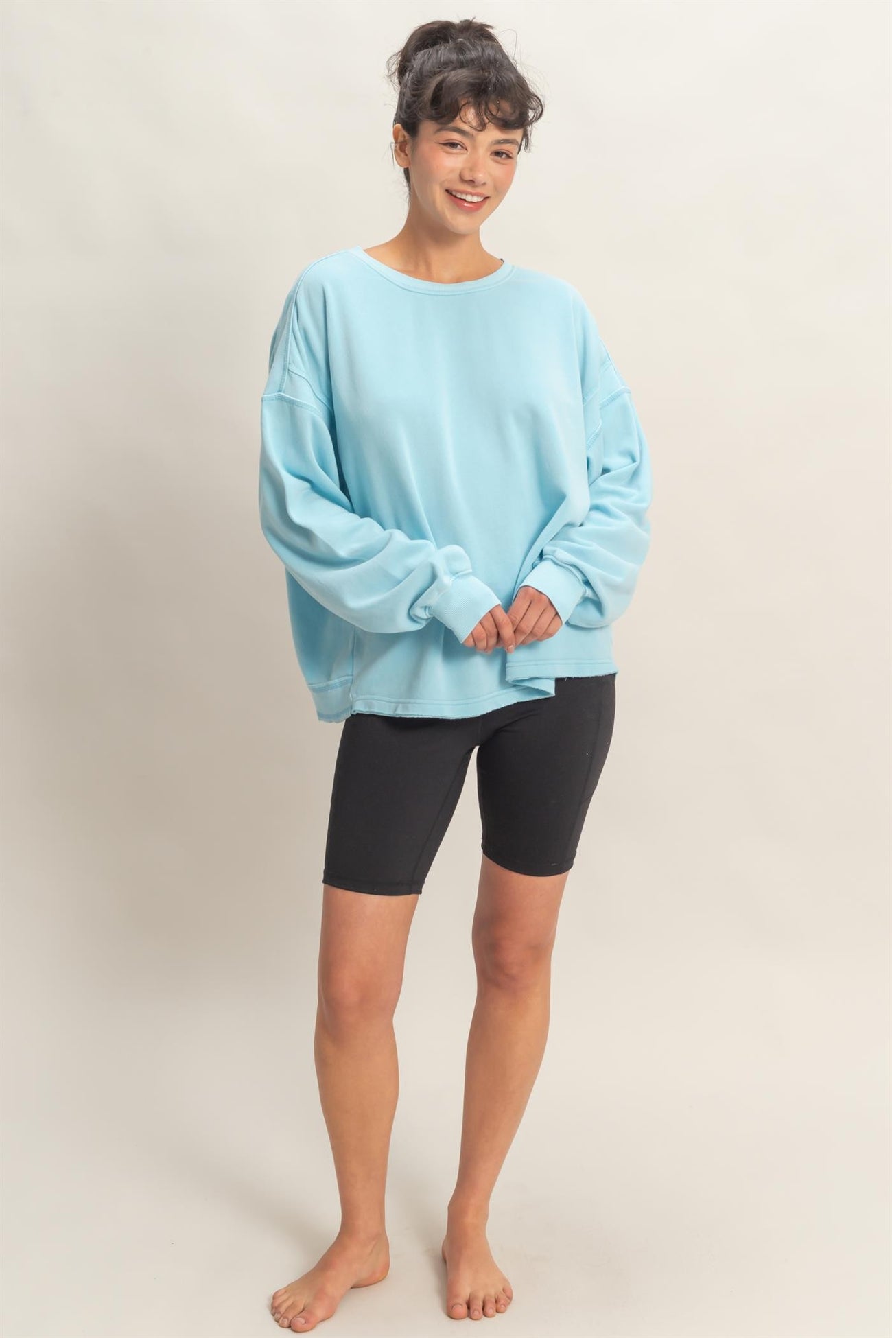 woman wearing a light blue oversized sweatshirt and black biker shorts, standing barefoot and smiling