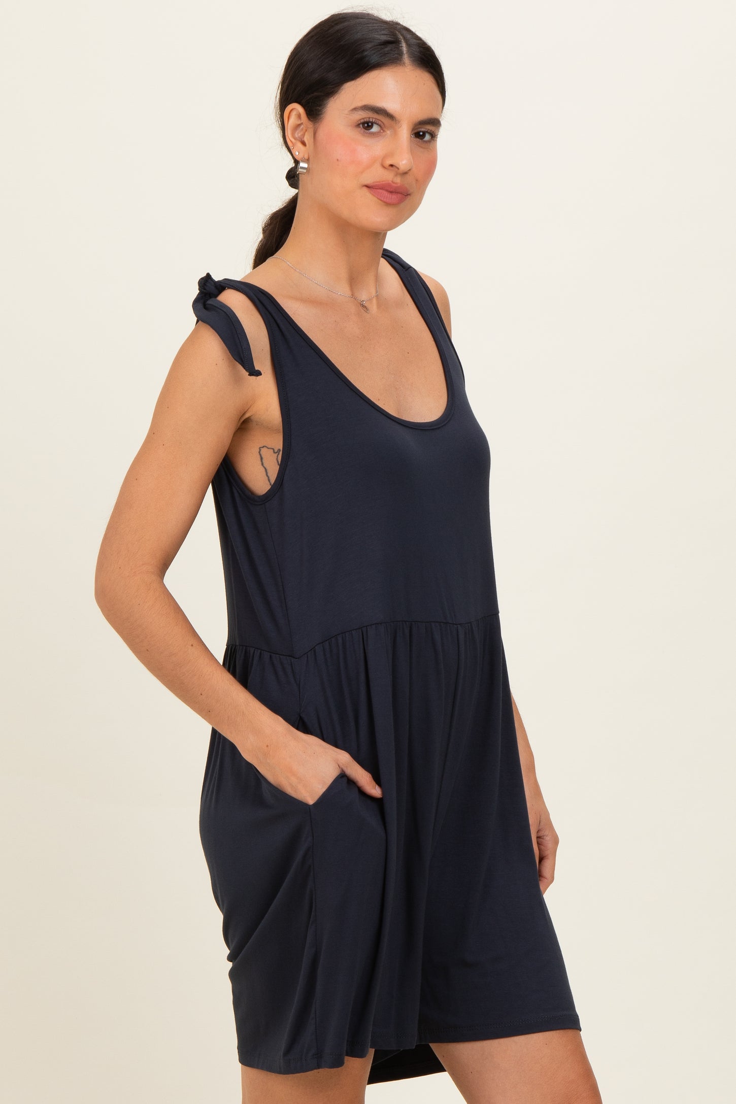 woman wearing a navy blue sleeveless dress with a gathered waist and side pockets, featuring tie detail on the straps
