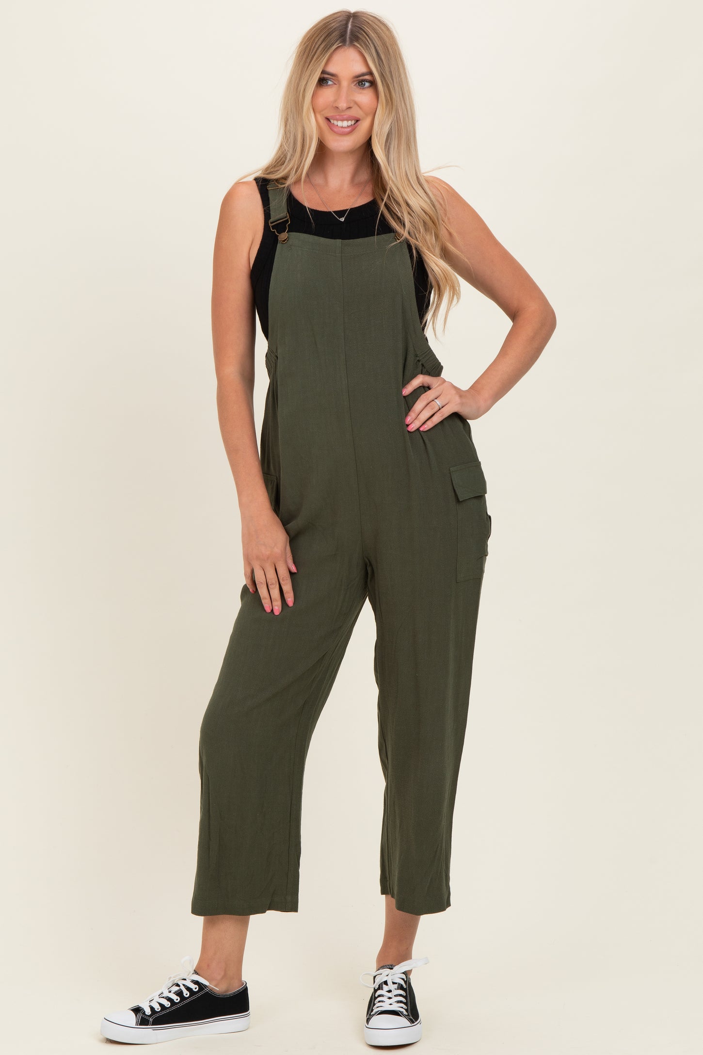 woman wearing an olive green sleeveless overall jumpsuit with side pockets and black sneakers