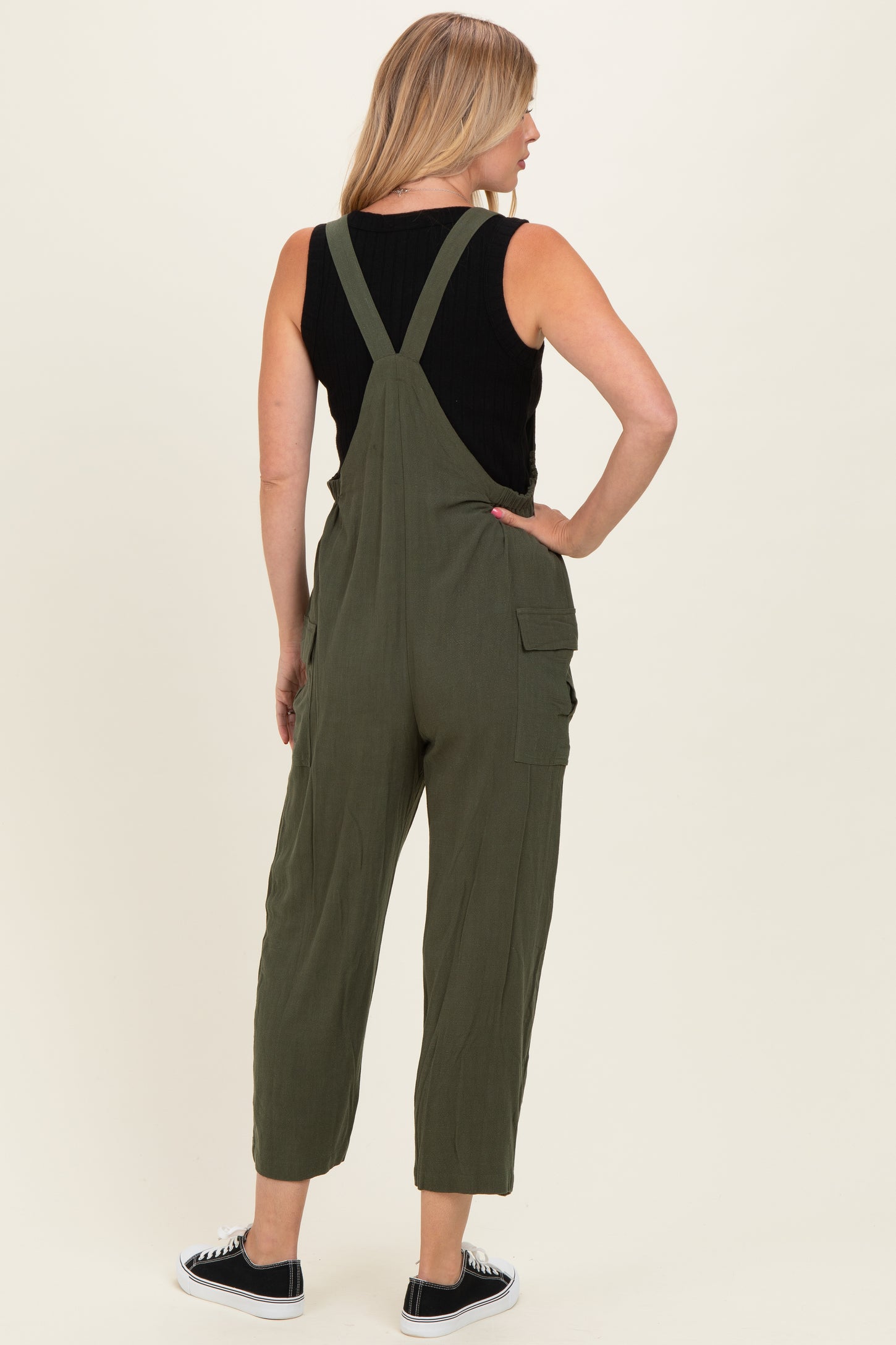 woman wearing an olive green overall jumpsuit with adjustable straps and side pockets, seen from the back