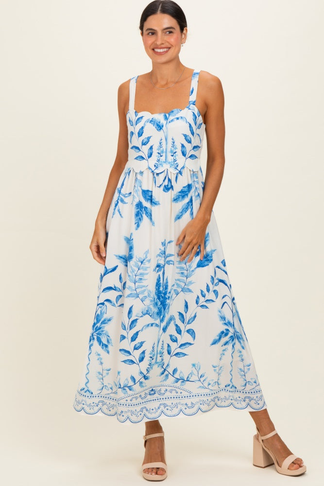woman wearing a white maxi dress with blue floral print and scalloped hem, standing against a neutral background