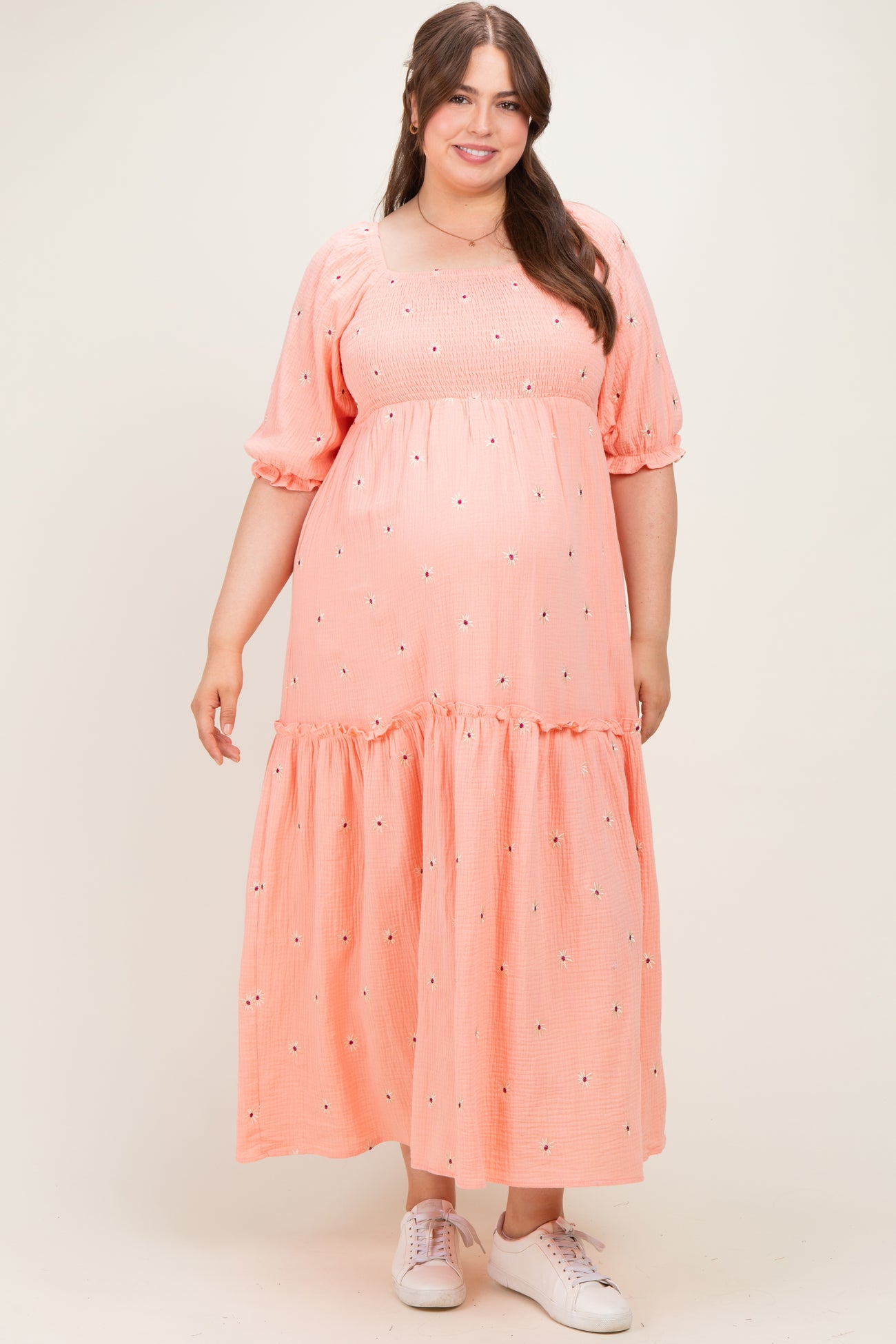 woman wearing a coral pink maternity plus maxi dress with puff sleeves and embroidered details, featuring a baby bump