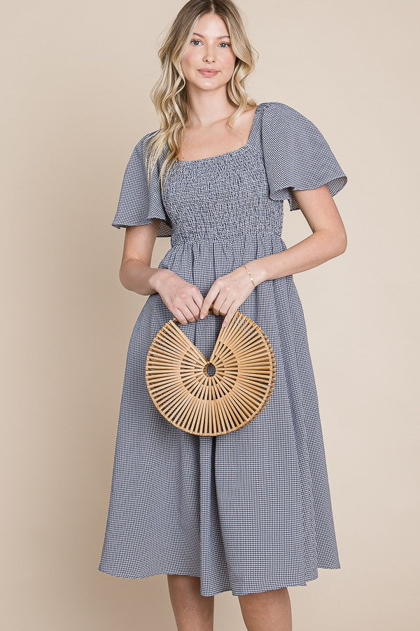 woman wearing a blue and white gingham smocked dress with short puffy sleeves and holding a bamboo handbag