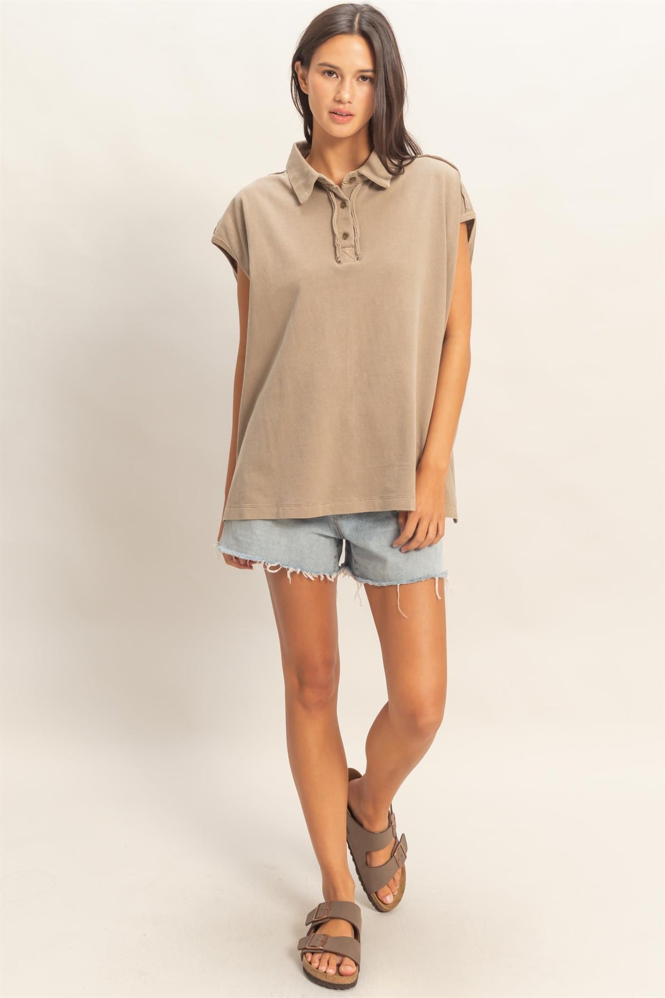 woman wearing a taupe short sleeve polo shirt and denim shorts, standing with one foot forward