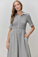 woman wearing a black and white gingham button-up dress with short sleeves and a smocked bodice, standing with one hand in her pocket