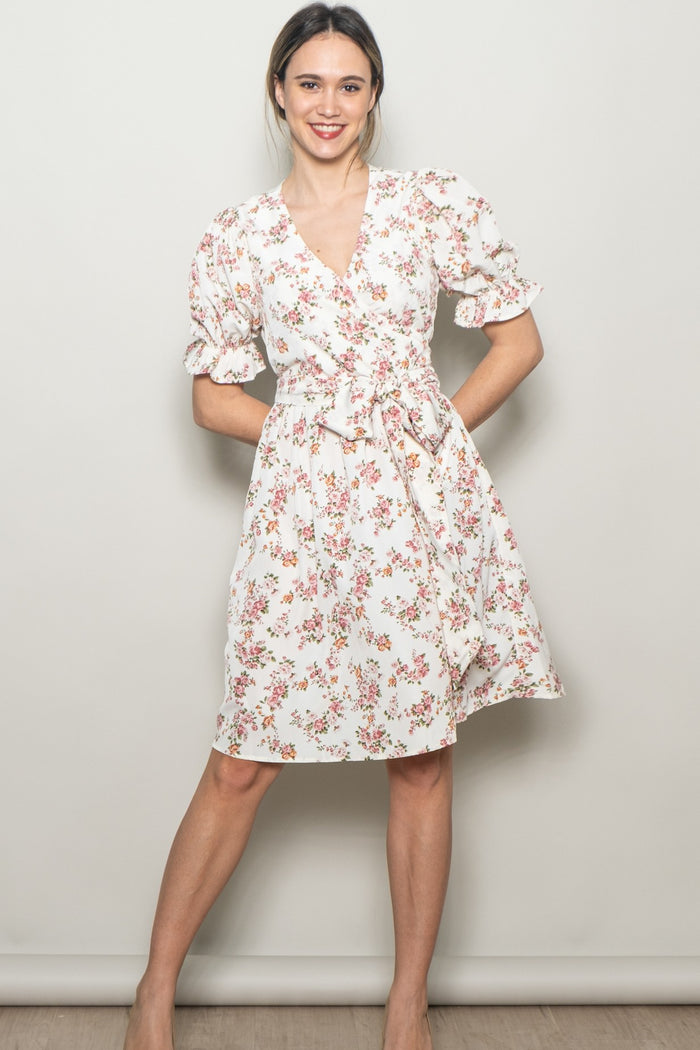 woman wearing a white floral wrap dress with puff sleeves and a tie waist, standing against a neutral background