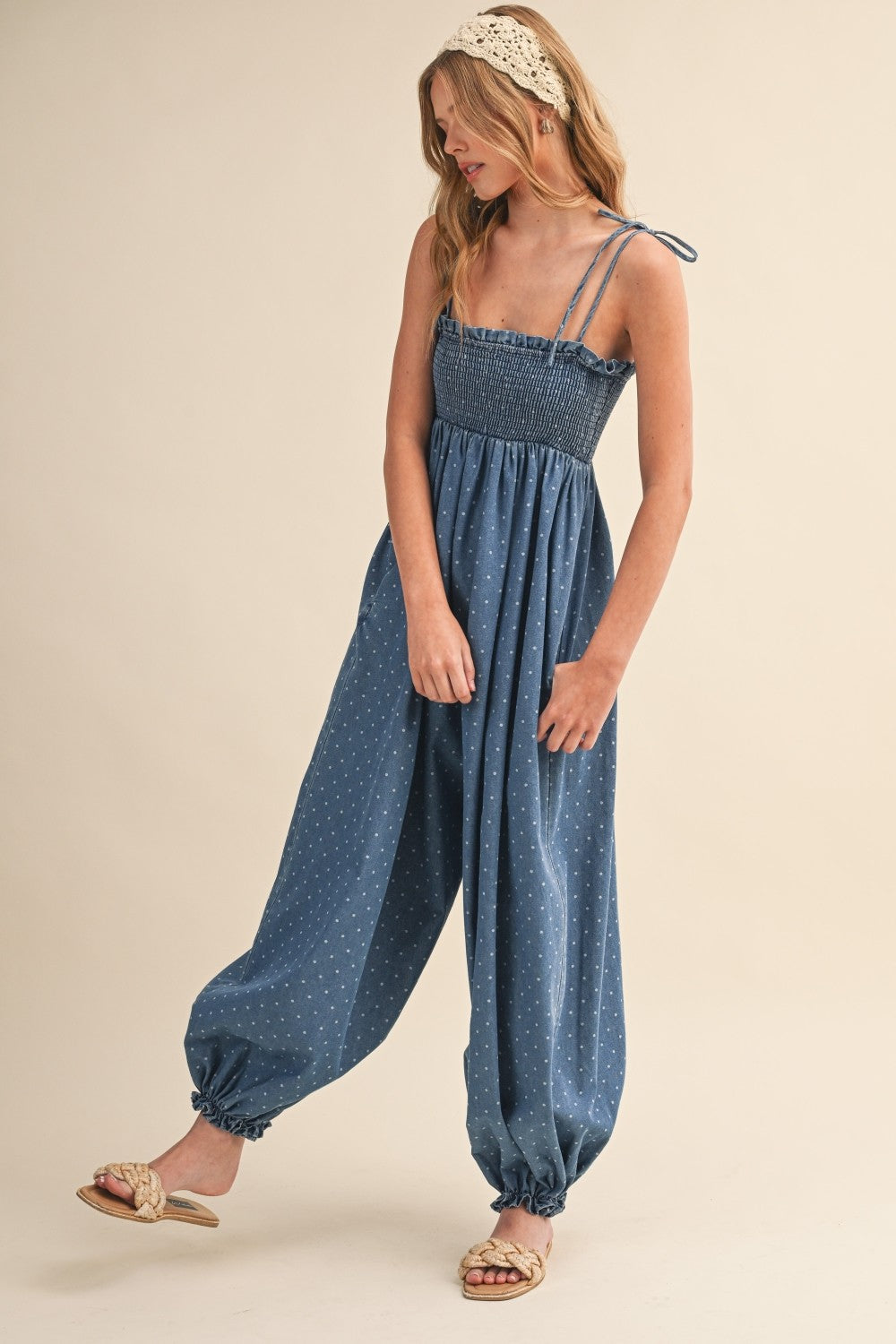woman wearing a blue smocked jumpsuit with polka dots and adjustable straps, styled with flat sandals and a headband