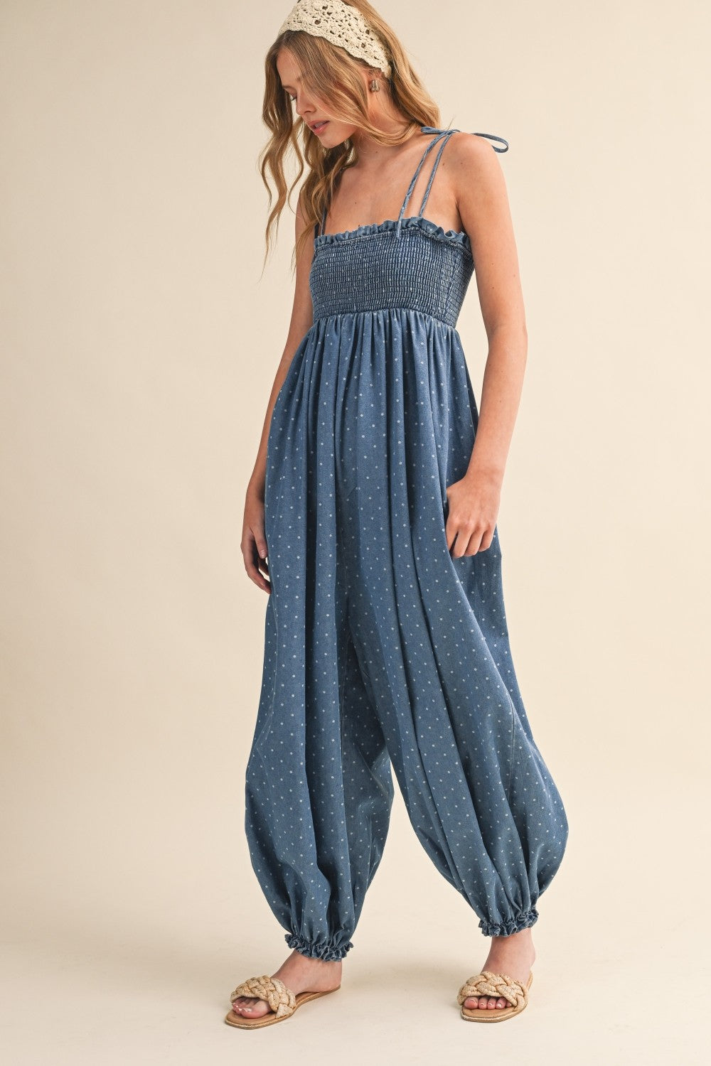 woman wearing a blue smocked jumpsuit with adjustable straps and gathered pants, styled with a headband and sandals