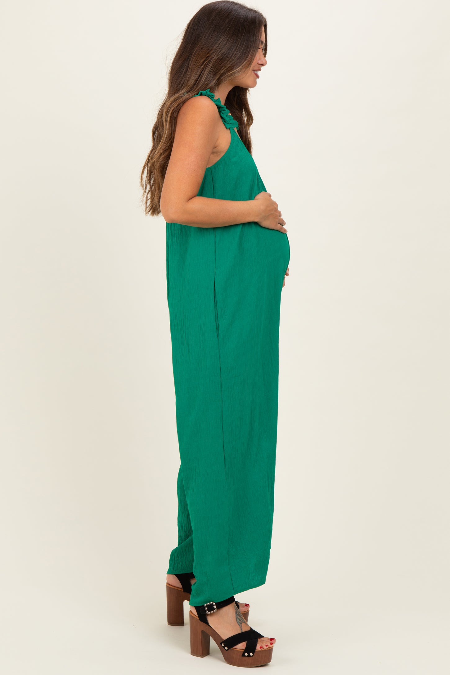 woman wearing a green sleeveless maternity jumpsuit, embracing her baby bump while standing sideways