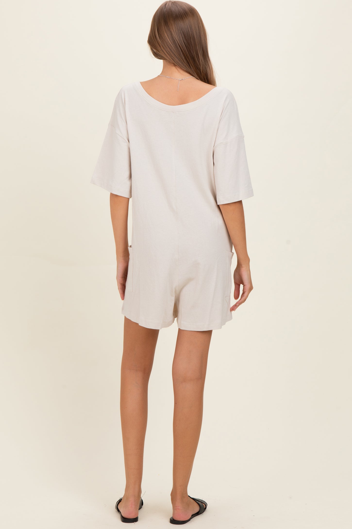 woman wearing a light beige short sleeve maternity romper, showing a relaxed fit and back view