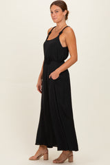 woman wearing a black sleeveless jumpsuit with a floral belt detail and pockets, paired with tan block-heeled sandals