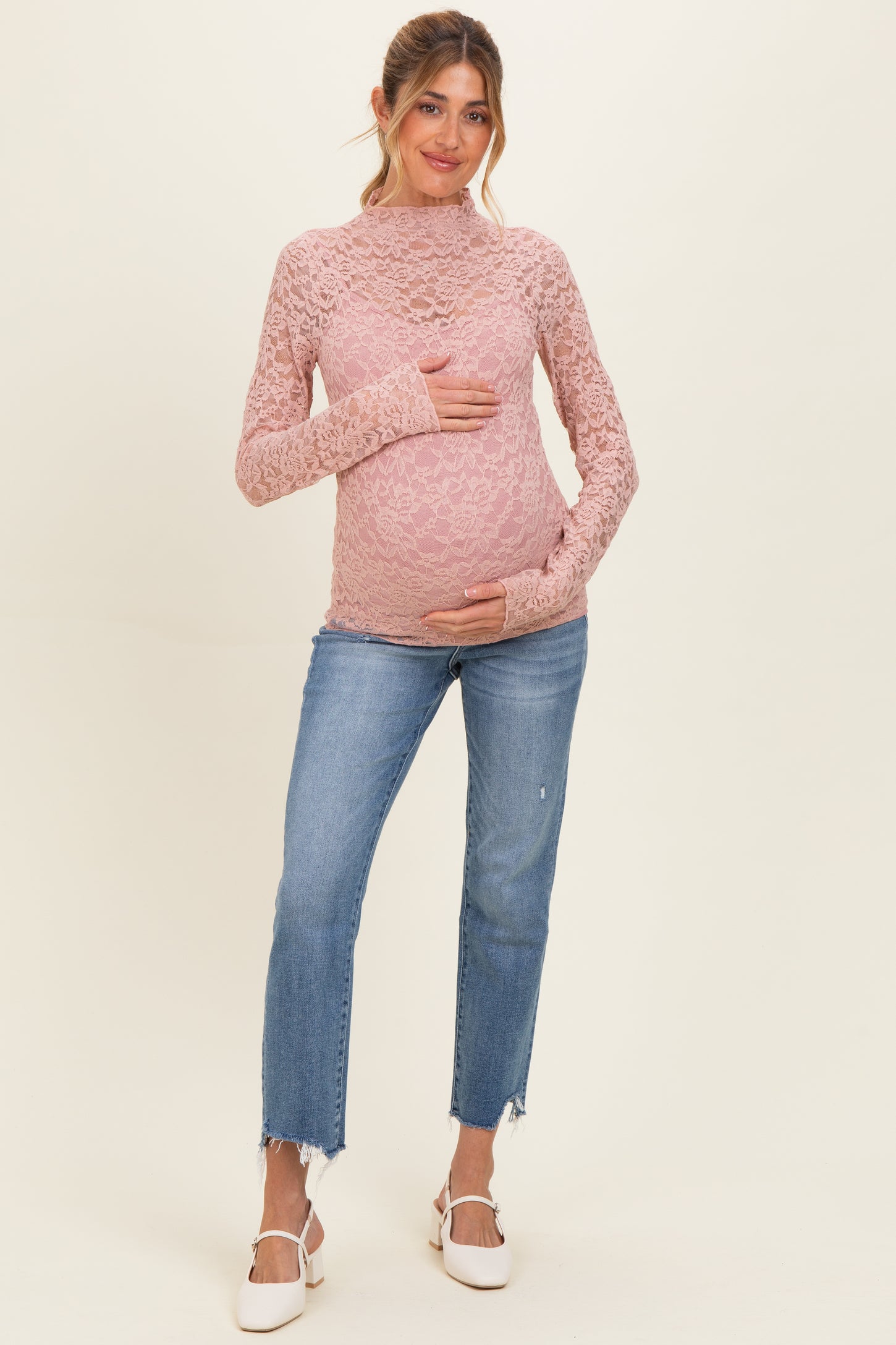 woman wearing a pink lace long sleeve maternity top and holding her baby bump with distressed blue jeans