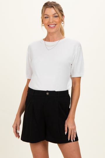 woman wearing a white short sleeve top and black high-waisted shorts, smiling and showing a casual maternity style