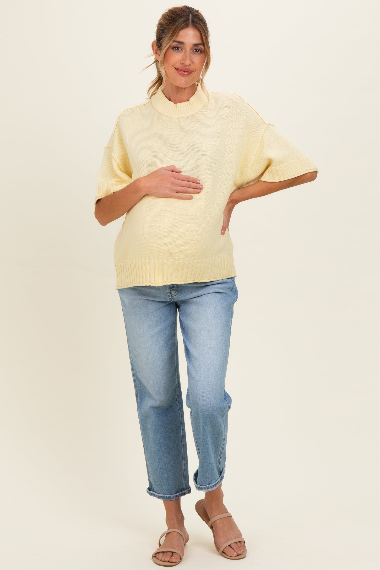 woman wearing a light yellow maternity sweater and blue jeans while holding her baby bump