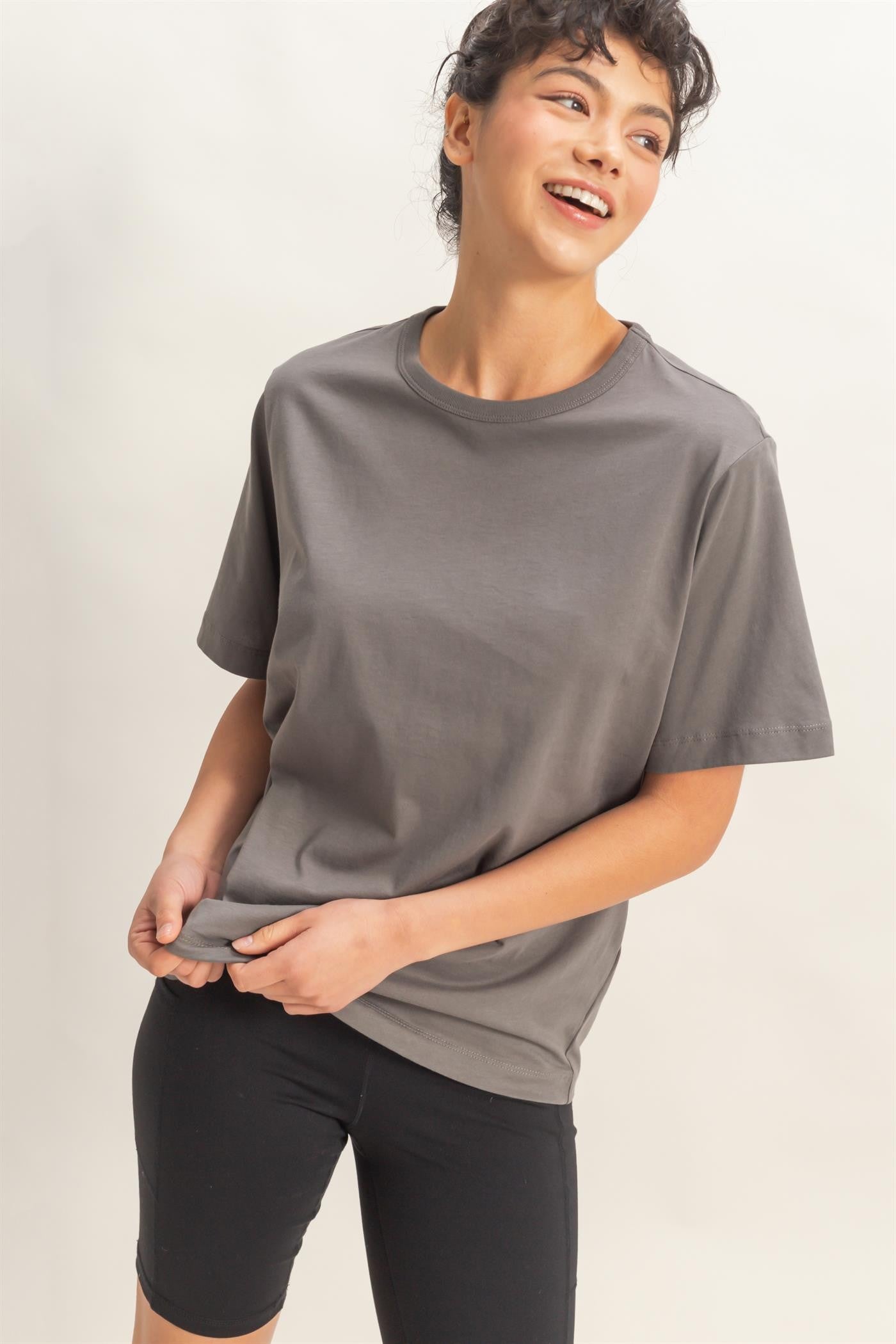 woman wearing a gray short sleeve t-shirt and black bike shorts, smiling while holding the t-shirt hem