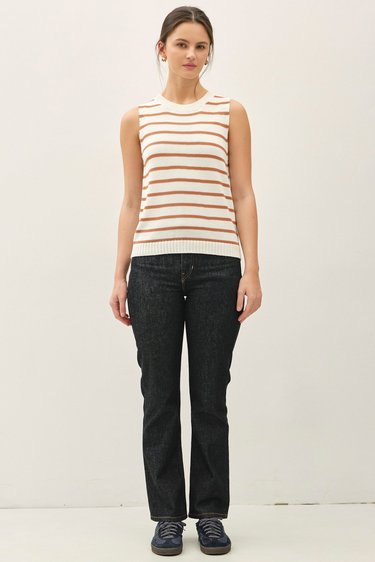 woman wearing a cream and orange striped sleeveless sweater with dark wash flared jeans and navy sneakers