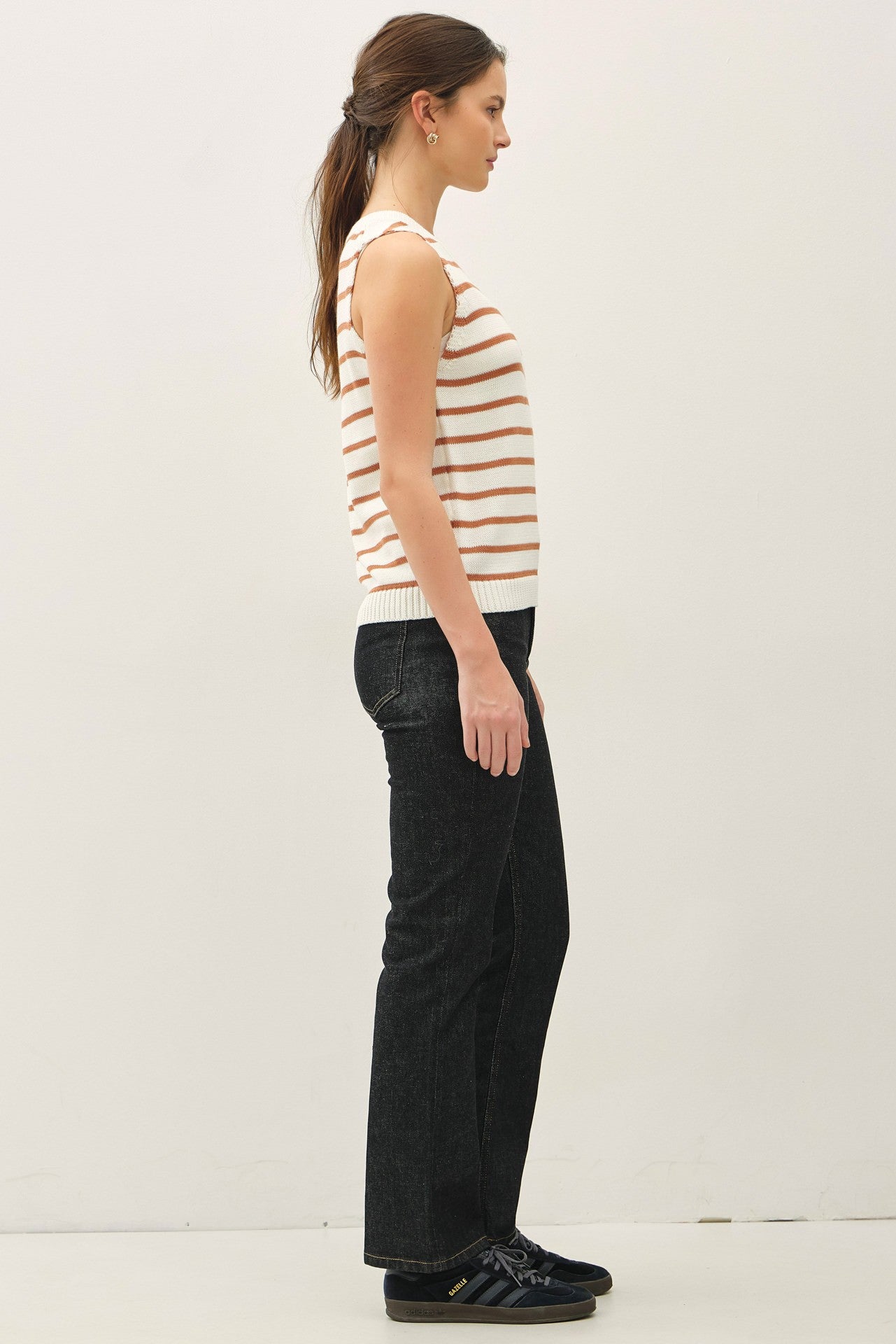 woman wearing a cream and rust striped sleeveless top and dark flared jeans, standing sideways against a plain background