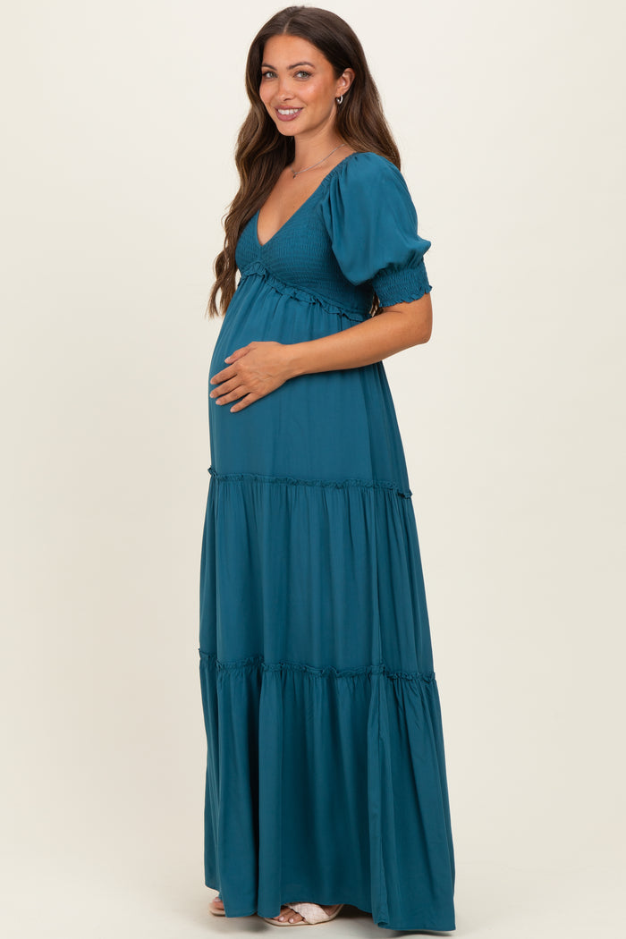 woman wearing a teal maternity maxi dress with puff sleeves and a tiered skirt, gently cradling her baby bump