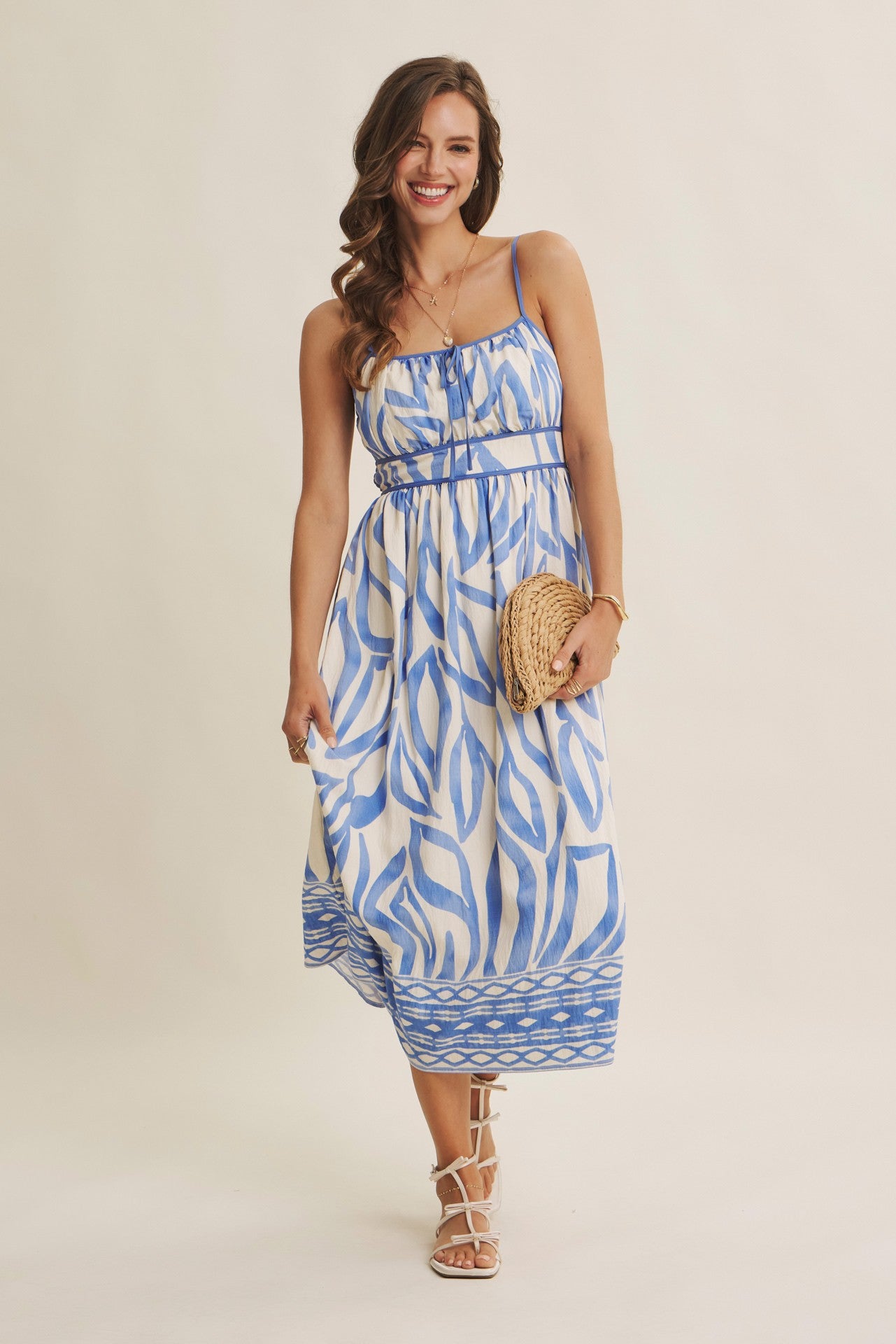 woman wearing a blue and white patterned midi dress with spaghetti straps and a belted waist, holding a straw clutch