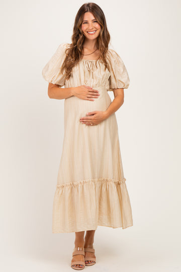 woman wearing a beige maternity maxi dress with puffed sleeves and a ruffled hem, gently holding her baby bump