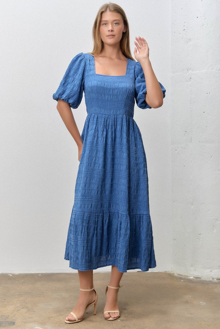 woman wearing a blue midi dress with square neckline and puff sleeves, standing against a white wall