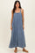 woman wearing a blue denim maxi dress with thin straps and side pockets, standing against a neutral background