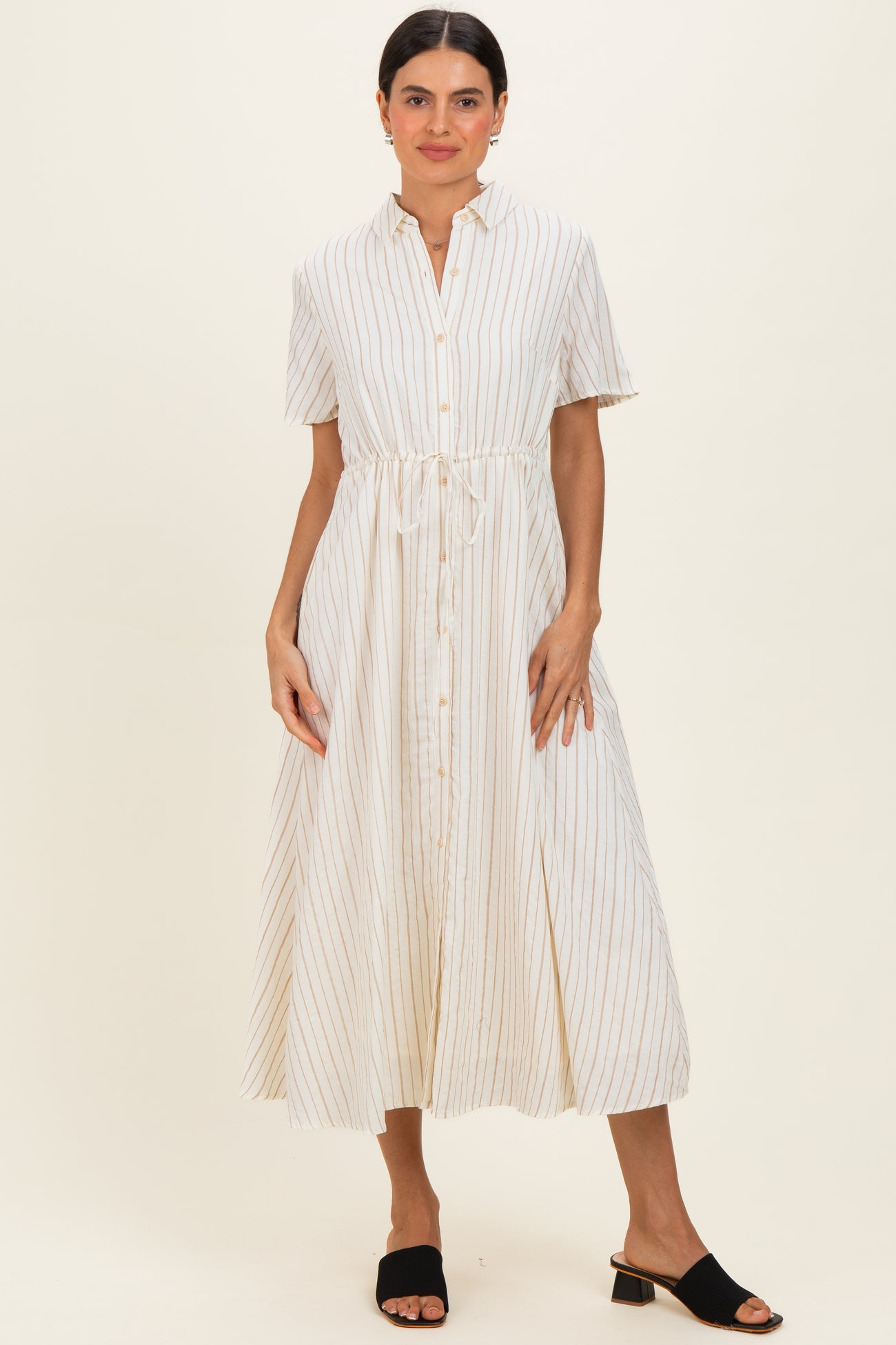 woman wearing a cream striped midi dress with short sleeves and a drawstring waist, paired with black slide sandals