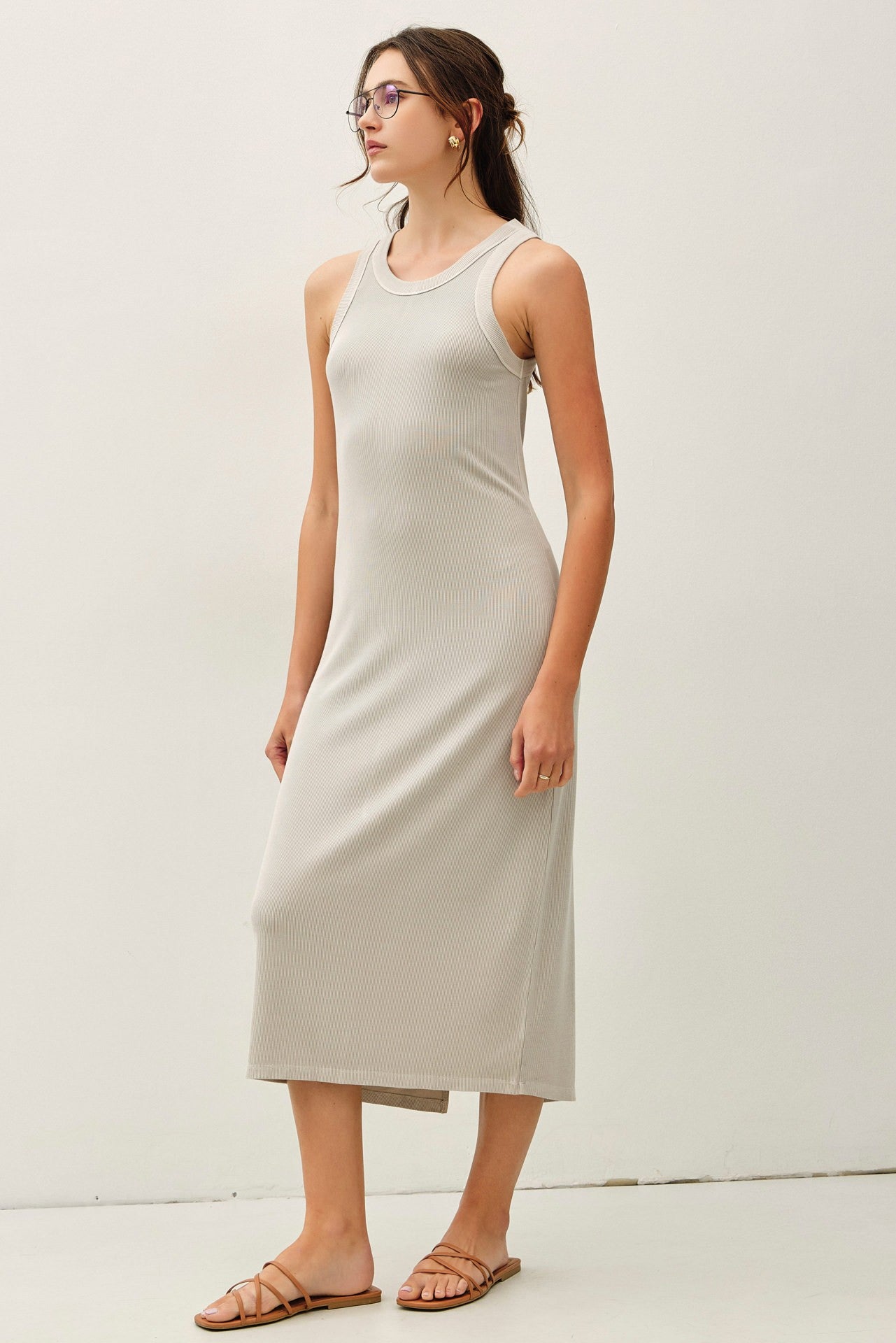 woman wearing a light beige sleeveless maxi dress with a fitted silhouette and side slit, standing against a plain background